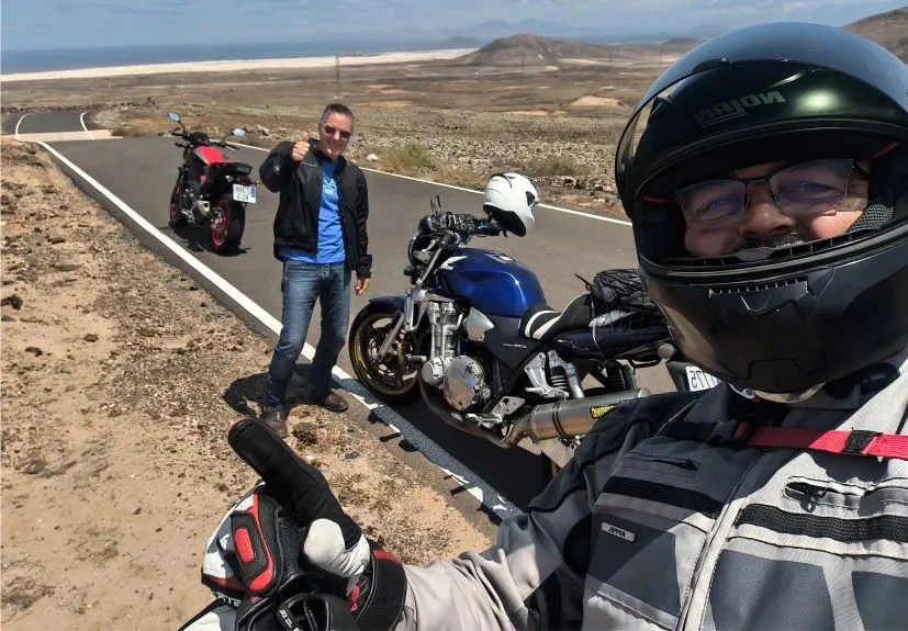 Motorcycle tour guide with a happy client stopped on a scenic secondary road in Fuerteventura, overlooking the Corralejo dunes and volcanic lava landscape.
